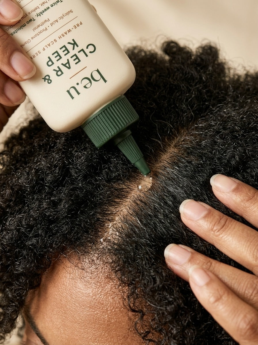 A close-up of a person applying a beige pre-wash soap and seal from the brand be.u to a section of dark curly hair.
