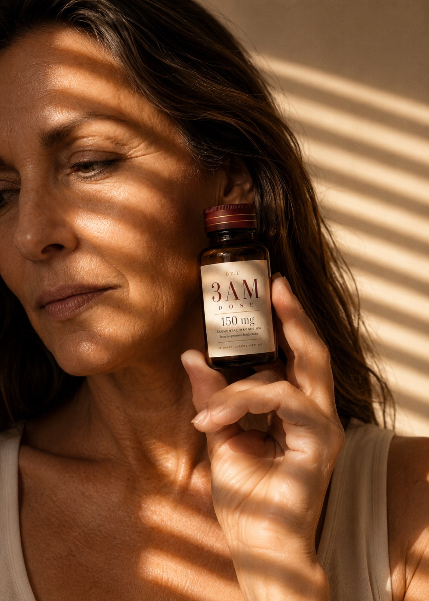 A woman in a beige tank top holds a small amber bottle labeled '3 AM' and '150 mg' near her face, with warm sunlight and window blinds casting shadows across her face and the bottle.